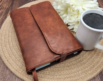 May include: A brown leather journal with a flap closure and a ribbon bookmark. The journal is placed on a woven mat, next to a white mug filled with a dark liquid and a white flower.