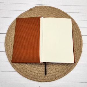 May include: A brown ribbed notebook with a blank white page open. The notebook is lying on a woven straw mat.