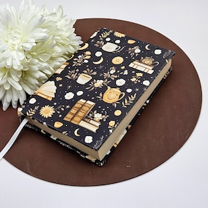 Pode incluir: Um livro de capa dura com uma capa preta com um padrão repetido de xícaras de chá, bules, livros, flores e luas em dourado e branco. Uma fita branca como marcador de livros é visível. O livro repousa sobre uma superfície circular marrom.