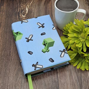 May include: A light blue book cover with pixelated green Creeper and sword patterns. It has a green ribbon bookmark. A white mug with dark liquid and green flowers are in the background.