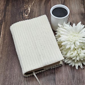 May include: A cream-colored corduroy book cover with a matching ribbon bookmark. A white mug filled with dark liquid and a white chrysanthemum flower are next to the book on a dark wood surface.