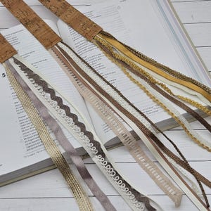 May include: A selection of cork and ribbon bookmarks with decorative ribbons and lace. The bookmarks are displayed on the open pages of a book. The ribbons are in shades of brown, beige, and white, with some gold accents. The cork tops are rectangular.