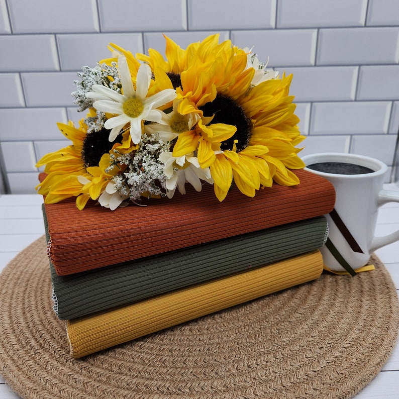 May include: A stack of four fabric swatches in shades of orange, green, and yellow. The fabric has a ribbed texture. A bouquet of sunflowers and white daisies sits on top of the fabric.