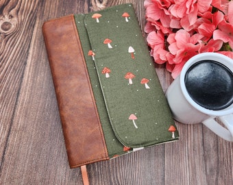 May include: A rectangular book cover featuring a dark olive green fabric panel with a repeating pattern of small red and white mushrooms and a brown faux leather spine. A thin peach-colored ribbon bookmark extends from the bottom edge of the cover.