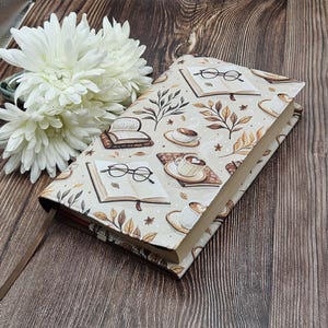 May include: A rectangular book cover featuring a cream-colored fabric with a repeating pattern of open books, black-rimmed eyeglasses, steaming coffee cups on saucers, and brown autumn leaves. A thin brown ribbon bookmark extends from the bottom.