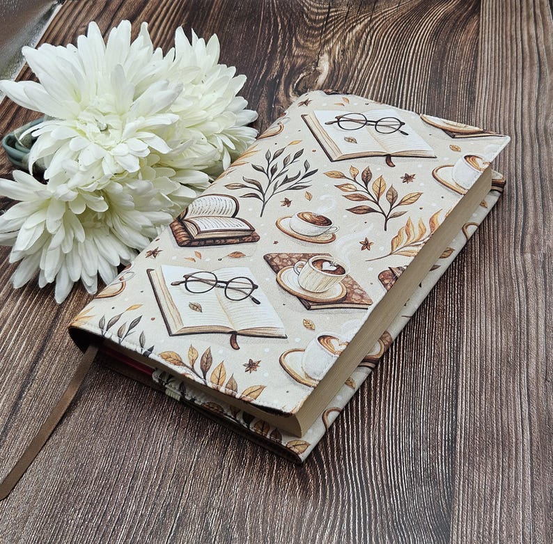 May include: A rectangular book cover featuring a cream-colored fabric with a repeating pattern of open books, black-rimmed eyeglasses, steaming coffee cups on saucers, and brown autumn leaves. A thin brown ribbon bookmark extends from the bottom.