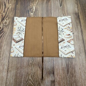 May include: A fabric book sleeve with a solid brown interior and a matching brown ribbon bookmark. The exterior flaps feature a cream-colored pattern of open books, round eyeglasses, coffee cups, and botanical leaf illustrations in brown tones.
