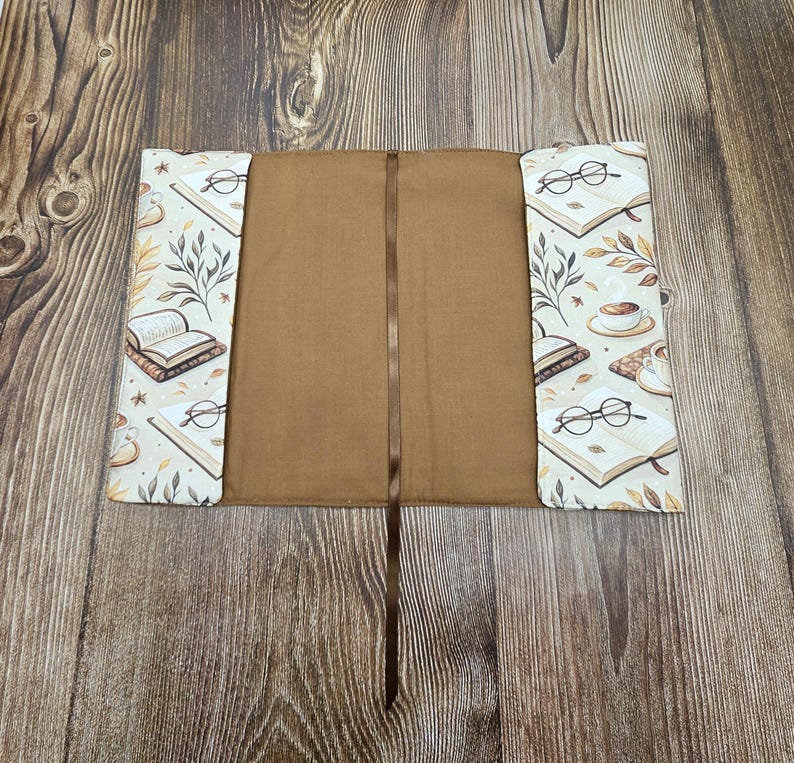 May include: A fabric book sleeve with a solid brown interior and a matching brown ribbon bookmark. The exterior flaps feature a cream-colored pattern of open books, round eyeglasses, coffee cups, and botanical leaf illustrations in brown tones.