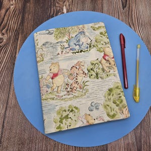 May include: A notebook with a cover depicting Winnie the Pooh and friends in a forest setting. The illustration includes Pooh Bear, Tigger, and Eeyore. A red pen and a gold pen are placed beside the notebook on a blue surface. The notebook is likely for writing.