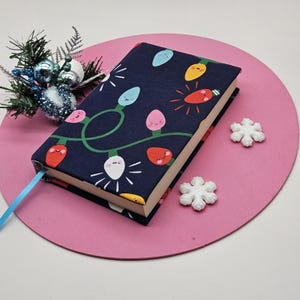 Puede incluir: Un libro de tapa dura con una cubierta azul marino que presenta una cadena de luces navideñas de colores con caras sonrientes. Una cinta de color azul claro sirve de marcapáginas y sobresale del libro. El libro descansa sobre una superficie circular rosa, con copos de nieve decorativos y una pequeña decoración navideña.