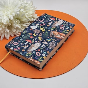 May include: A hardback journal with a dark blue cover featuring a whimsical forest design. The cover showcases illustrations of owls, foxes, mushrooms, and various flowers and birds. A yellow ribbon bookmark is visible. The journal rests on an orange circular mat.
