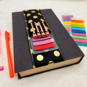 Puede incluir: Una libreta negra con lunares dorados y un marcapáginas de tela negra y dorada. El marcapáginas tiene cinco clips metálicos dorados y un bolsillo de plástico transparente con notas adhesivas de colores.