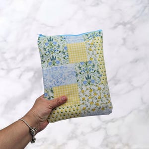 May include: A patchwork zippered pouch with a light blue zipper. The pouch features a mix of floral, gingham, and patterned fabrics in shades of blue, yellow, and white. The design has a quilted appearance.