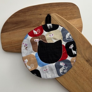 May include: A round, quilted pot holder with a black cat pocket and a loop for hanging. The fabric features a repeating pattern of cat silhouettes in various colors. The pot holder rests on two wooden cutting boards.