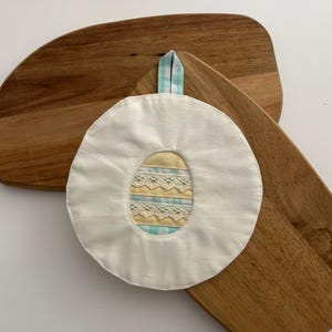 Könnte beinhalten: Ein weißer, runder Topflappen mit einem eiförmigen Design mit gelben, blauen und Spitzendetails. Er hat eine blau-weiß karierte Schlaufe zum Aufhängen. Der Topflappen liegt auf zwei Holzschneidebrettern.