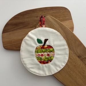 May include: A white, round pot holder with an apple design, featuring a green leaf and brown stem. The apple's interior is filled with colorful fabric. The pot holder has a red and white loop for hanging. Two wooden cutting boards are in the background.