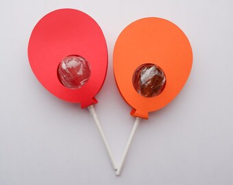 May include: Two lollipop treats shaped like red and orange balloons. The lollipops are on white sticks and have clear wrappers.