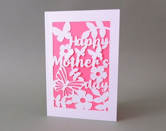 May include: White card with a pink background and a cut-out design of flowers, butterflies, and leaves. The text "Happy Mother's Day" is cut out of the card.