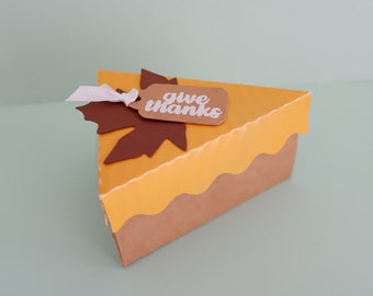 May include: A brown and yellow pie-shaped box with a brown leaf and a tag that says "give thanks".