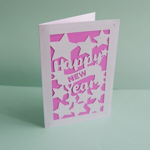 May include: A white card with a pink background and a cut-out design of stars and the words "Happy New Year".