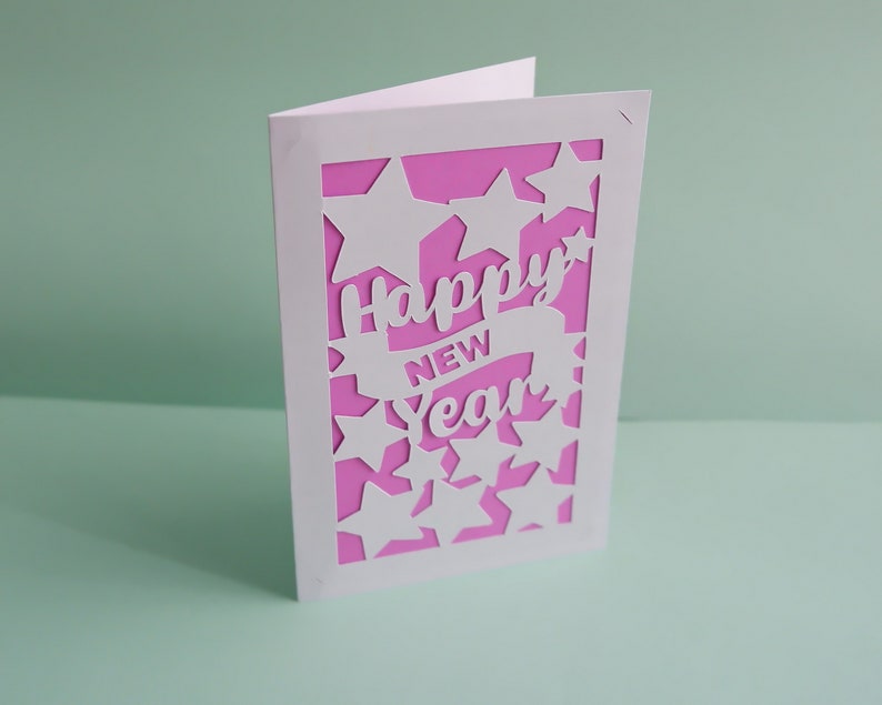 May include: A white card with a pink background and a cut-out design of stars and the words "Happy New Year".