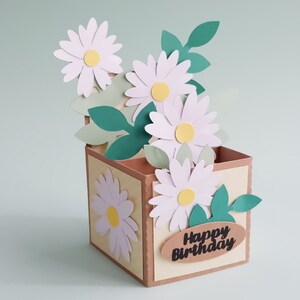 May include: A brown paper box with a pop-up card design featuring white daisies and green leaves. The box has a "Happy Birthday" message on the front.