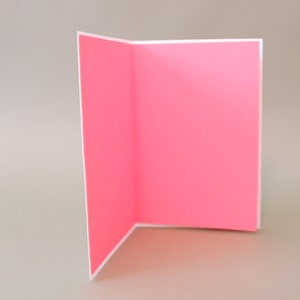 Puede incluir: Una tarjeta en blanco con un interior rosa y un exterior blanco. La tarjeta est&aacute; doblada por la mitad y est&aacute; de pie sobre una superficie gris claro.