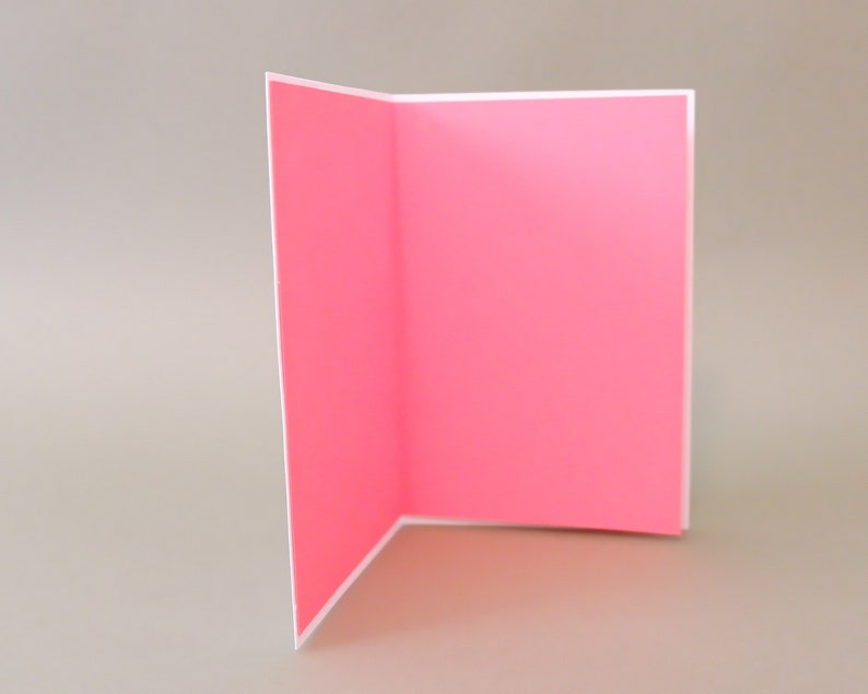Puede incluir: Una tarjeta en blanco con un interior rosa y un exterior blanco. La tarjeta est&aacute; doblada por la mitad y est&aacute; de pie sobre una superficie gris claro.