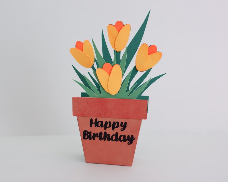K&ouml;nnte beinhalten: Eine Papierbastelkarte mit einer Topfpflanze mit gelben und orangefarbenen Tulpen und gr&uuml;nen Bl&auml;ttern. Der Topf ist braun und tr&auml;gt den Text "Happy Birthday".