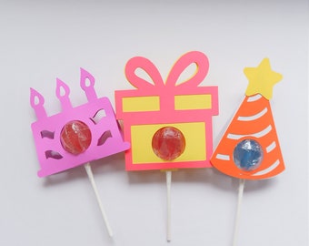 May include: Three colorful paper lollipop holders shaped like a birthday cake, a gift box, and a party hat. Each holder has a round hole to insert a lollipop.
