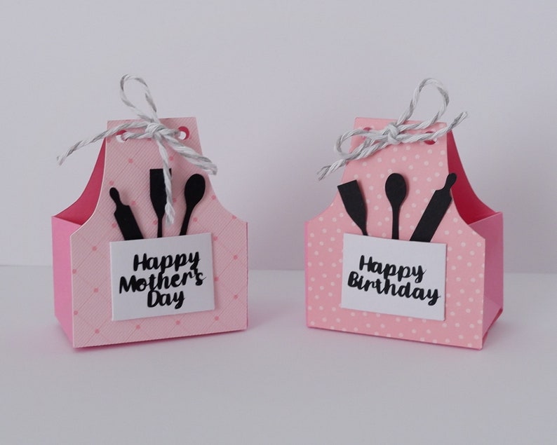 May include: Two pink paper gift boxes shaped like aprons with white polka dots. The boxes have black silhouettes of kitchen utensils and a white tag with the text "Happy Mother's Day" and "Happy Birthday".