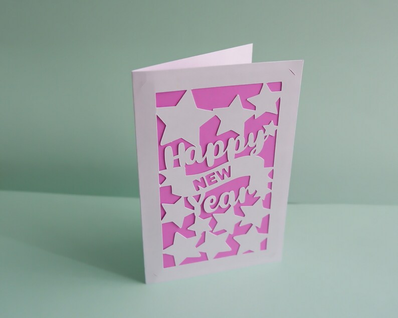 May include: A white card with a pink background and a cut-out design of stars and the words "Happy New Year".