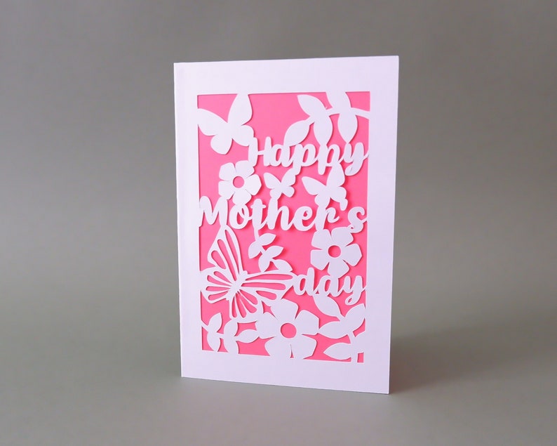 Puede incluir: Tarjeta blanca con fondo rosa y un dise&ntilde;o recortado de flores, mariposas y hojas. El texto "Happy Mother's Day" est&aacute; recortado de la tarjeta.