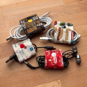 May include: Four fabric cord organizers with snap buttons, each wrapped around a coiled cable. One features a city grid pattern, one has New York themed graphics with the word Broadway, one shows London icons, and one displays white crowns on red.