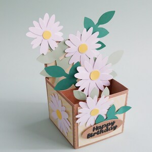 May include: A handmade pop-up card with a brown box and a bouquet of white daisies with yellow centers. The box has a yellow and white striped pattern and the text "Happy Birthday" on a brown oval tag.