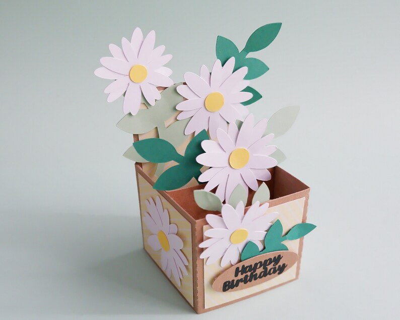 May include: A handmade pop-up card with a brown box and a bouquet of white daisies with yellow centers. The box has a yellow and white striped pattern and the text "Happy Birthday" on a brown oval tag.