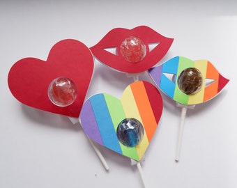 May include: Four colorful paper lollipop holders with cutouts for lollipops. The holders are shaped like a heart, lips, and a rainbow heart. Each holder has a lollipop inside.