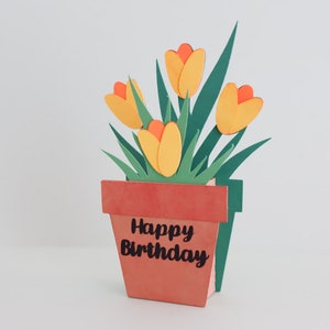 K&ouml;nnte beinhalten: Eine handgefertigte Papierbastelkarte mit einem Pop-up-Design, das eine Topfpflanze mit gelben und orangefarbenen Bl&uuml;ten und gr&uuml;nen Bl&auml;ttern zeigt. Der Topf ist braun und tr&auml;gt den Text "Happy Birthday".