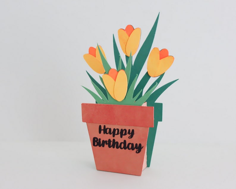 K&ouml;nnte beinhalten: Eine handgefertigte Papierbastelkarte mit einem Pop-up-Design, das eine Topfpflanze mit gelben und orangefarbenen Bl&uuml;ten und gr&uuml;nen Bl&auml;ttern zeigt. Der Topf ist braun und tr&auml;gt den Text "Happy Birthday".