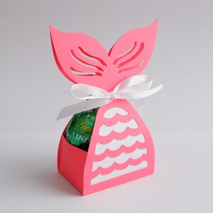 May include: A pink paper mermaid tail-shaped gift box with a white ribbon. The box has a cut-out design and holds a green and gold wrapped chocolate candy. The box is on a white background.