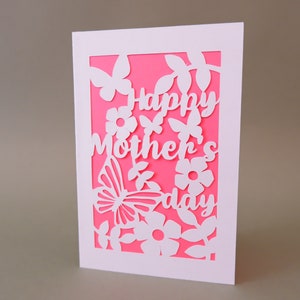 Puede incluir: Una tarjeta blanca con fondo rosa y un dise&ntilde;o recortado en blanco de flores, mariposas y el texto "Happy Mother's Day".