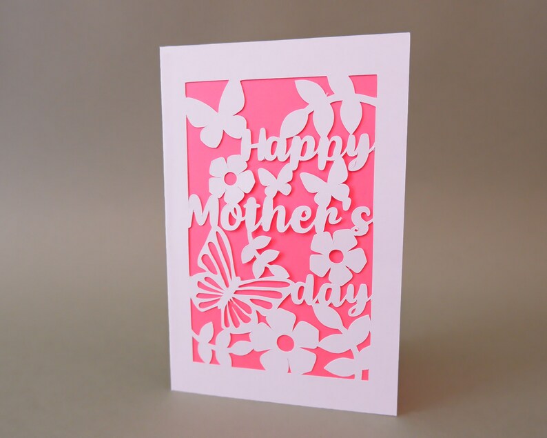 Puede incluir: Una tarjeta blanca con fondo rosa y un dise&ntilde;o recortado en blanco de flores, mariposas y el texto "Happy Mother's Day".