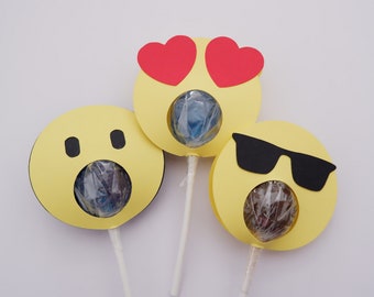 May include: Three yellow paper lollipop wrappers with different emoji faces. The first wrapper has a heart-eyed emoji, the second has a surprised emoji, and the third has a sunglasses emoji. Each wrapper has a hole in the center to hold a lollipop.