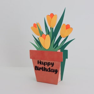 K&ouml;nnte beinhalten: Eine Papierbastelkarte mit einem Pop-up-Blumenarrangement in einem Terrakotta-Topf. Die Blumen sind gelb mit orangefarbenen Zentren und gr&uuml;nen Bl&auml;ttern. Der Topf tr&auml;gt die Aufschrift "Happy Birthday".