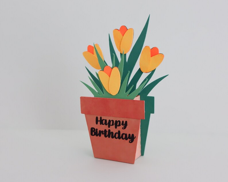 K&ouml;nnte beinhalten: Eine Papierbastelkarte mit einem Pop-up-Blumenarrangement in einem Terrakotta-Topf. Die Blumen sind gelb mit orangefarbenen Zentren und gr&uuml;nen Bl&auml;ttern. Der Topf tr&auml;gt die Aufschrift "Happy Birthday".