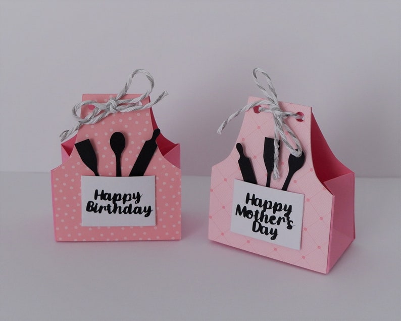 May include: Two pink paper gift boxes shaped like aprons with black kitchen utensil cutouts. The box on the left says "Happy Birthday" and the box on the right says "Happy Mother's Day".