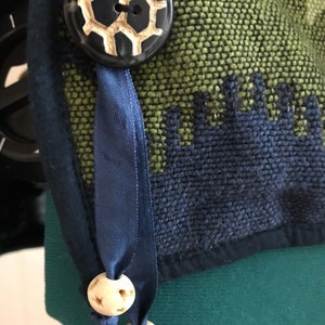 May include: A close-up of a blue ribbon tied with two white bone beads. The ribbon is attached to a green and blue fabric with a black button with a white design.