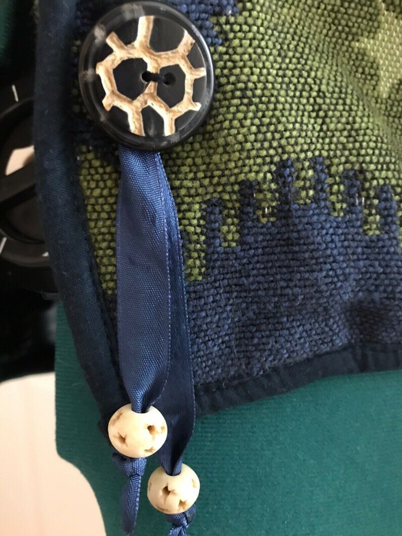 May include: A close-up of a blue ribbon tied with two white bone beads. The ribbon is attached to a green and blue fabric with a black button with a white design.