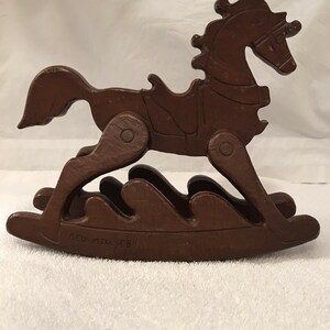 以下が含まれることがあります： 鞍と鐙が付いた茶色の木製のロッキングホース。馬は、彫刻された波模様の台座の上で揺れています。台座には「Red Mill © 69」と刻印されています。
