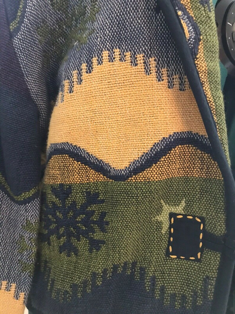 May include: A blue, green, and yellow woven jacket with a snowflake and star design. The jacket has a black trim and a patch on the sleeve.